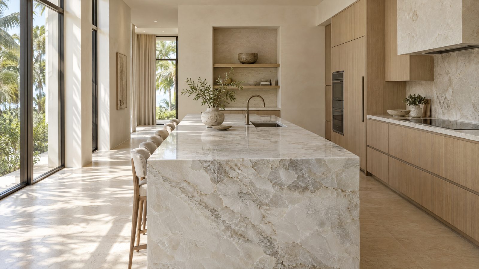 Long kitchen island topped with white and grey quartzite with irregular veining, white-oak cabinets and tall windows with palm shadows